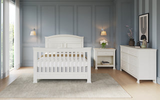 A stylish bedroom with a white bed, nightstand, dresser, and blue paneled walls. Flowers, art, and lights decorate the space, while the Child Craft Westgate 4-in-1 Convertible Crib in Vintage Linen adds charm. A beige rug covers the wooden floor.