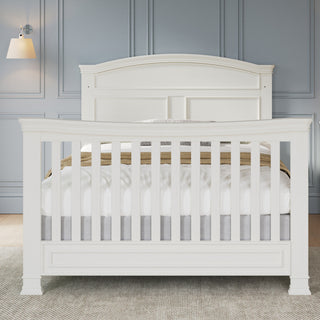The Child Craft Westgate 4-in-1 Convertible Crib in Vintage Linen, featuring vertical slats, adjustable mattress support, and decorative paneling, stands on a light rug in a room with gray paneled walls and a wall-mounted lamp.