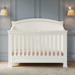 The Child Craft Westgate 4-in-1 Convertible Crib in Vintage Linen stands against a blue paneled wall, flanked by modern sconces with beige shades. A light wood floor and gray rug complete this serene nursery setting.