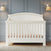 The Child Craft Westgate 4-in-1 Convertible Crib in Vintage Linen stands against a blue paneled wall, flanked by modern sconces with beige shades. A light wood floor and gray rug complete this serene nursery setting.