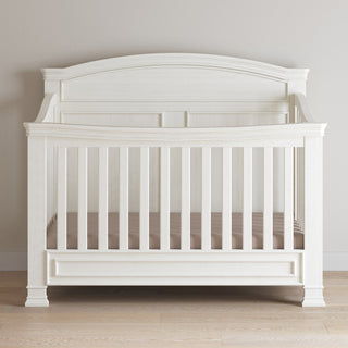 The Child Craft Westgate 4-in-1 Convertible Crib in Vintage Linen features vertical slats, a curved headboard, a light brown mattress, and adjustable mattress support, standing on a light wood floor against a beige wall.