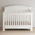 The Child Craft Westgate 4-in-1 Convertible Crib in Vintage Linen features vertical slats, a curved headboard, a light brown mattress, and adjustable mattress support, standing on a light wood floor against a beige wall.