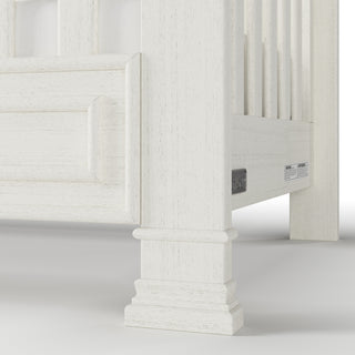 Close-up of the Child Craft Westgate 4-in-1 Convertible Crib in Vintage Linen, showing decorative leg and side details. Labels and the adjustable mattress support are visible beneath the crib frame, set against a plain background.