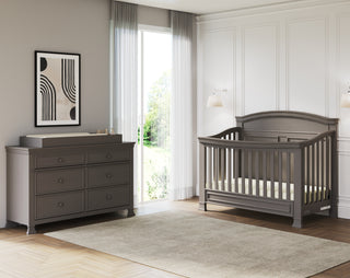 A modern nursery featuring a gray crib, Child Craft Westgate Double Dresser in Chelsea Gray with a changing pad, abstract wall art, a soft beige rug, and a large window with sheer curtains bathing the room in natural light.