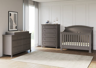A modern nursery with gray furniture, including a crib, a changing table, and the Child Craft Westgate 5-Drawer Chest in Chelsea Gray, set on a light rug over hardwood floors with white paneled walls and sunlight from a large window.