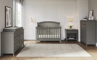A nursery features the Child Craft Westgate 4-in-1 Convertible Crib in Chelsea Grey with matching gray dresser, chest, and nightstand on a light rug over wood floors. White paneled walls, two wall lamps, a window, and modern art complete the space.