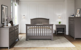 A stylish nursery features a gray crib on a beige rug, complemented by the Child Craft Westgate 5-Drawer Chest in Chelsea Gray, white paneled walls, modern decor, and soft natural light from a nearby window.