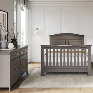A stylish nursery features a gray crib and the Child Craft Westgate Double Dresser in Chelsea Gray, with artwork, decorative vases, white paneled walls, curtained window, wooden floor, and a beige rug for a serene finish.