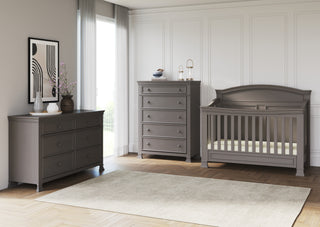 Modern nursery featuring the Child Craft Westgate 4-in-1 Convertible Crib in Chelsea Grey, tall and wide dressers, a neutral rug on herringbone wood floors, white paneled walls, large window with sheer curtains, framed art, and floral vases.