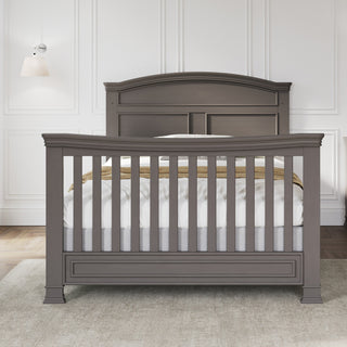 The Child Craft Westgate 4-in-1 Convertible Crib in Chelsea Grey stands against a white paneled wall on a light rug, styled with white bedding and a beige blanket for a cozy, versatile nursery look.