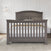 The Child Craft Westgate 4-in-1 Convertible Crib in Chelsea Grey stands against a white paneled wall on a light rug, styled with white bedding and a beige blanket for a cozy, versatile nursery look.