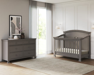 A modern nursery features the Child Craft Westgate Double Dresser in Chelsea Gray and a matching crib, neutral walls, a large window with sheer curtains, a beige rug on wood floors, minimalist art, and a flower vase on the dresser.