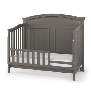 The Child Craft Westgate 4-in-1 Convertible Crib in Chelsea Grey features a high, arched headboard, slatted sides, and an open side for easy access, shown with a white mattress. Designed to grow with your child.