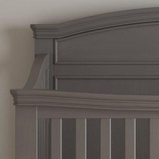 Close-up of the Child Craft Westgate 4-in-1 Convertible Crib in Chelsea Grey, featuring a dark pine wood headboard with decorative moldings and vertical slats, blending timeless design and lasting durability against a light wall.