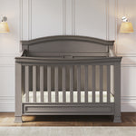 The Child Craft Westgate 4-in-1 Convertible Crib in Chelsea Grey, featuring an arched headboard, stands on a hardwood floor against white paneled walls, with two beige-shaded wall-mounted lamps above.