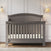 The Child Craft Westgate 4-in-1 Convertible Crib in Chelsea Grey, featuring an arched headboard, stands on a hardwood floor against white paneled walls, with two beige-shaded wall-mounted lamps above.