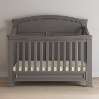 The Child Craft Westgate 4-in-1 Convertible Crib in Chelsea Grey, featuring vertical slats and a curved headboard, is placed on a light wood floor with a white and beige striped sheet against a plain light-colored wall.