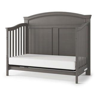 The Child Craft Westgate 4-in-1 Convertible Crib in Chelsea Grey features an arched headboard, solid panel back, slatted sides, white mattress, and adjustable support for toddler bed use.