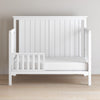 The Child Craft Toddler Guard Rail in Matte White features sturdy wood construction, slatted sides, and a left partial safety rail, shown on a light wood floor against a plain beige wall.