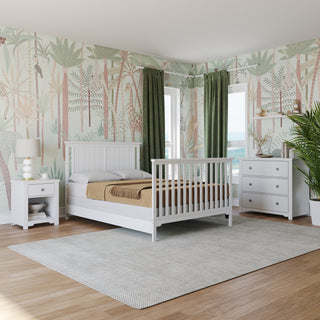 A bright bedroom features a white bed with Child Craft F06401 Full-Size Bed Rails in matte white, plus a nightstand, dresser, green curtains, large plant, tropical wallpaper, wood floors with a gray rug, and sunlight streaming through corner windows.