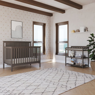 A modern nursery with a dark wood crib, Child Craft’s Flat Top Baby Changing Table in Dapper Gray for storage, patterned wallpaper, two large windows, a geometric rug, a potted plant, and shelves with decor and baby essentials.