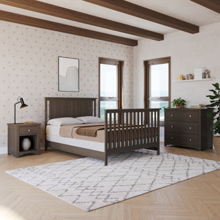 A modern bedroom features the Child Craft Harmony 3-Drawer Dresser with Changing Table Topper in Dapper Gray, a wooden bed and nightstand, large windows, light patterned walls, geometric rug, green plant, and framed abstract art.