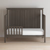 A dark wood toddler bed from Child Craft features strong construction, a high slatted headboard, the F09501 Dapper Gray Toddler Guard Rail, and a white mattress, set on a light wood floor against a plain wall.