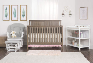 A baby nursery featuring a wooden crib, Child Craft Cozy Glider and Ottoman in matte white, stuffed lamb, white changing table with baby supplies, wall art, and soft rugs on a dark wood floor. Light walls and curtains add a calm atmosphere.