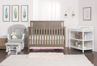 A cozy nursery features the Child Craft Harmony 33" Kids Toy Box in Dusty Heather, a wooden crib, gray glider with stuffed lamb, white changing table with baby supplies, framed leaf art, soft rugs on wood floor, and sunlight from the window.