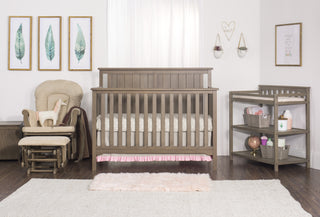 A cozy nursery with a wooden crib, changing table, beige rocker, soft rug, and the Child Craft Harmony 33" Kids Toy Box in Dusty Heather. Leaf print and heart wall art complement hanging plants; white curtains create a softly lit space.