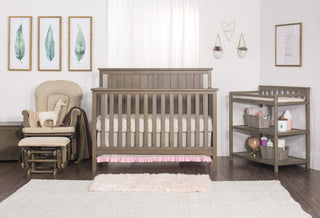 A cozy nursery featuring a wooden crib, Child Craft’s Cozy Glider and Ottoman in Dusty Heather, a changing table with baskets, framed leaf prints, a fluffy rug, and natural light streaming through white curtains.