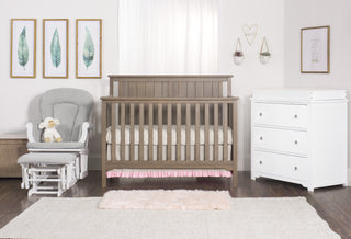 A nursery with the Child Craft Harmony 33" Kids Toy Box in Dusty Heather, a wooden crib, white dresser, gray rocking chair with a stuffed lamb, leafy wall art, heart-shaped hanging, and a fluffy rug on the wooden floor.