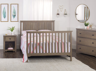 A cozy bedroom features a neatly made bed with a pink and white comforter. A Child Craft Harmony 3-Drawer Dresser with Changing Table Topper in Dusty Heather stands beside a nightstand with a plant, soft lighting, wall art, and hanging shelves.