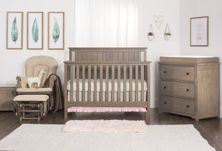 A cozy nursery features a wooden crib with pink bedding, a beige glider chair with a stuffed horse, framed leaf prints, soft rugs on wood floors, and the Child Craft Harmony 3-Drawer Dresser with Changing Table Topper in Dusty Heather.