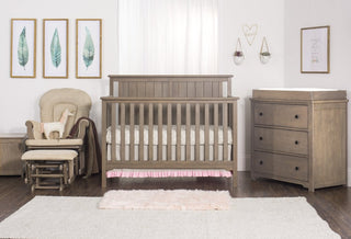 A cozy nursery features a wooden crib, matching dresser with changing pad, pink crib skirt, plush rug, and natural light. The Child Craft Cozy Glider and Ottoman in Dusty Heather offer comfy seating beside framed art and a curtained window.