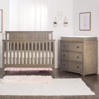A cozy nursery features a wooden crib, the Child Craft Harmony 3-Drawer Dresser with Changing Table Topper in Dusty Heather, a fluffy pink rug, white walls adorned with art and hanging planters, and sunlight through a window with white curtains.