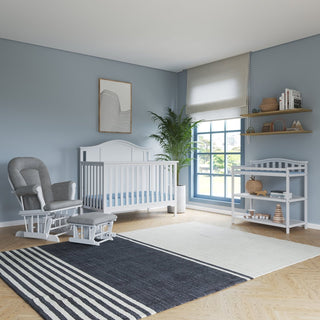 A modern nursery with blue walls features a white crib, Child Craft Cozy Glider and Ottoman in Matte White, a changing table, shelves with toys and books, large window with blinds, potted plant, and striped rug on wooden floors.