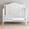 A Child Craft Toddler Guard Rail in Matte White, featuring sturdy wood construction and a modern curved design, is shown on a light wood floor against a beige wall. The partially open guard rail ensures both safety and style.