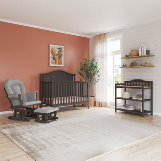 A modern nursery features stylish furnishings: a gray crib, Child Craft's Arch Top Baby Changing Table in Dapper Gray, a glider chair with ottoman, plus a peach accent wall, large rug, wood floors, shelves, and sheer curtains for a cozy touch.