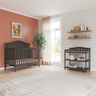 A cozy nursery includes a dark wood crib, Child Craft Arch Top Baby Changing Table in Dapper Gray with storage, peach accent wall, potted plant, decor shelves, and a large window with sheer curtains for natural light.