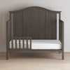 A Child Craft F09501 Toddler Guard Rail in Dapper Gray features strong wood construction, a partial safety rail, white mattress, and curved headboard, shown on a light wood floor against a plain light-colored wall.
