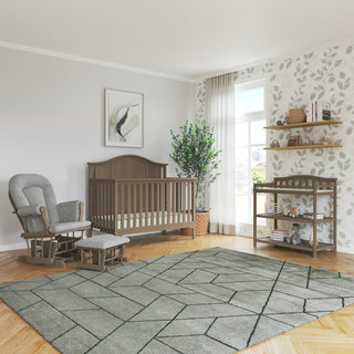 A cozy nursery with a wooden crib, Child Craft Cozy Glider and Ottoman in Dusty Heather, open shelving with books and toys, leafy wallpaper accent, large window with sheer curtains, potted plant, and patterned rug on wood floor.
