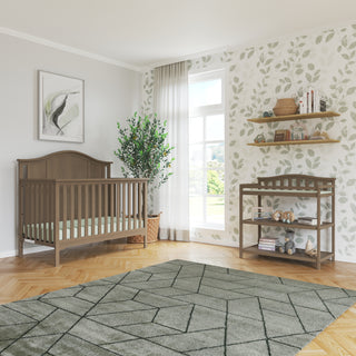 A cozy nursery featuring Child Craft's Arch Top Baby Changing Table in Dusty Heather, wooden furniture, leafy wallpaper, decor shelves, a potted plant, large window with sheer curtains, framed art, and a geometric-patterned rug on wood floors.