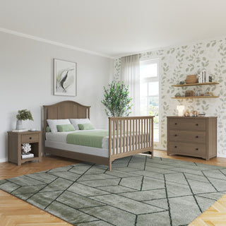 A cozy bedroom features a wooden bed, green and white bedding, nightstand, leafy wallpaper, large rug, potted plant, wall art, shelves with books and decor, and a Child Craft Harmony 3-Drawer Dresser with Changing Table Topper in Dusty Heather. Sunlight fills the room.