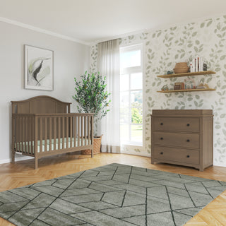 A cozy nursery features a wooden crib, green plant, leafy wallpaper, wall shelves with decor, framed art, sheer-curtained window, and a patterned rug. The Child Craft Harmony 3-Drawer Dresser with Changing Table Topper in Dusty Heather completes the space.