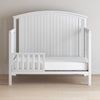 The Child Craft Toddler Guard Rail in Matte White features sturdy wood construction, a high slatted backrest, partial toddler guard rail, and a plain white mattress, set against a light wall on a wooden floor.