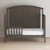 A Child Craft F09501 Toddler Guard Rail in dapper gray, featuring strong wood construction, a slatted half-rail guard, arched headboard, and white mattress is set on a light wood floor against a plain white wall.