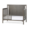 The Child Craft Toddler Guard Rail in Dapper Gray features strong wood construction, vertical slats on three sides, an open side for easy access, and comes with a white mattress.