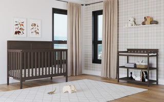 Modern nursery featuring a dark wood crib and the Child Craft Flat Top Baby Changing Table in Dapper Gray with baby changing pad, beige curtains, plaid accent wall, animal art, plush toys, books on shelf, light rug, and toy elephant on hardwood floor.