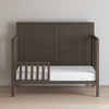 A Child Craft Toddler Guard Rail in dapper gray, crafted from sturdy wood with a paneled headboard, partial guard rail, and white mattress, sits against a light wall on a light wood floor.